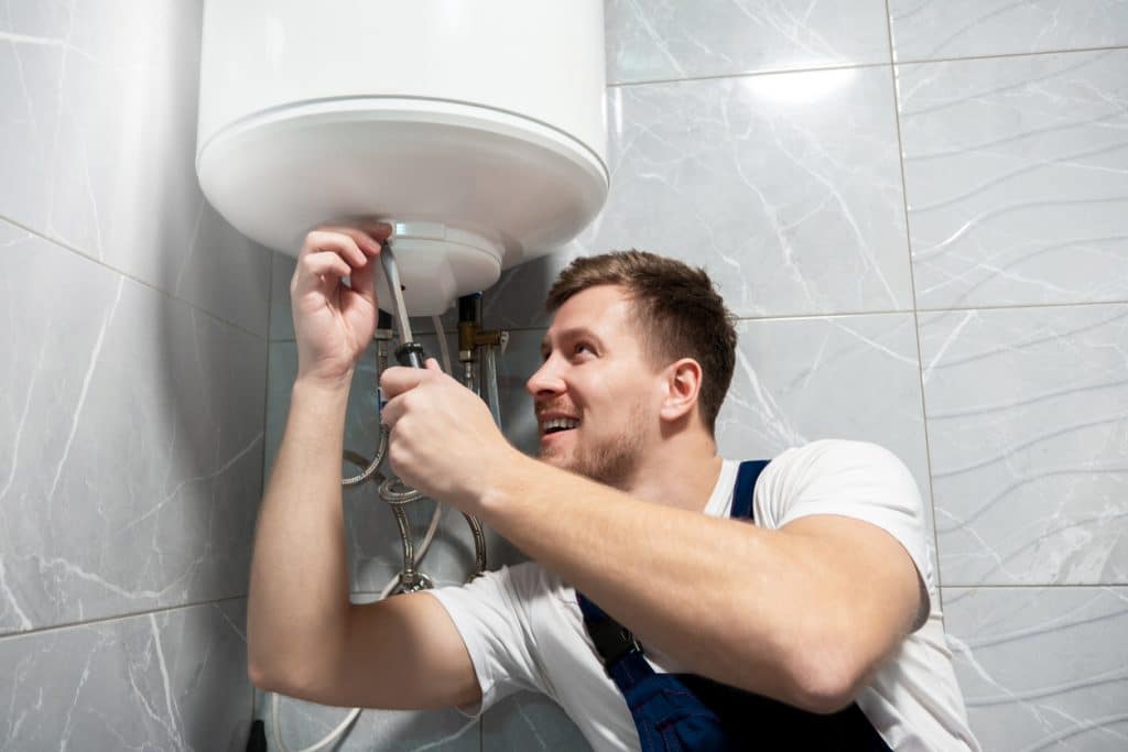 young handsome smiling man worker in uniform repairing water heater using screwdriver at home in the toilette professional repair service.